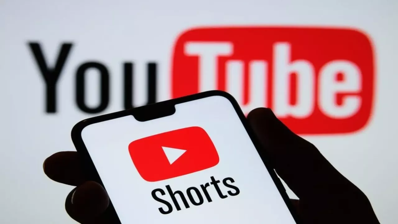 Akıllı telefonda açık olan YouTube Shorts uygulaması, arka planda büyük YouTube logosu ile görülüyor.